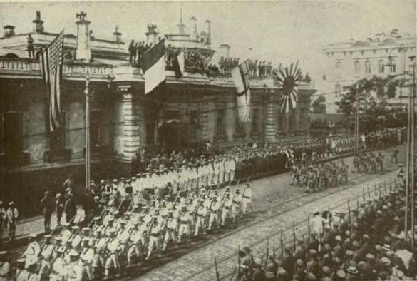 Groups of American, British, and Japanese soldiers parade down a street in front a building with America, French, British, and Japanese flags