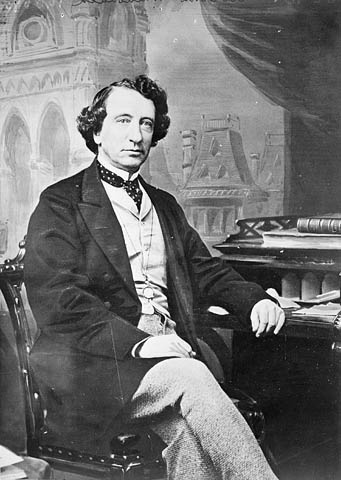 Seated photographic portrait of Sir John A. Macdonald in suit with arm resting on desk with papers