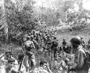 Large group of Marines, many wearing helmets, in various states of rest in a field surrounded by trees on Guadalcanal