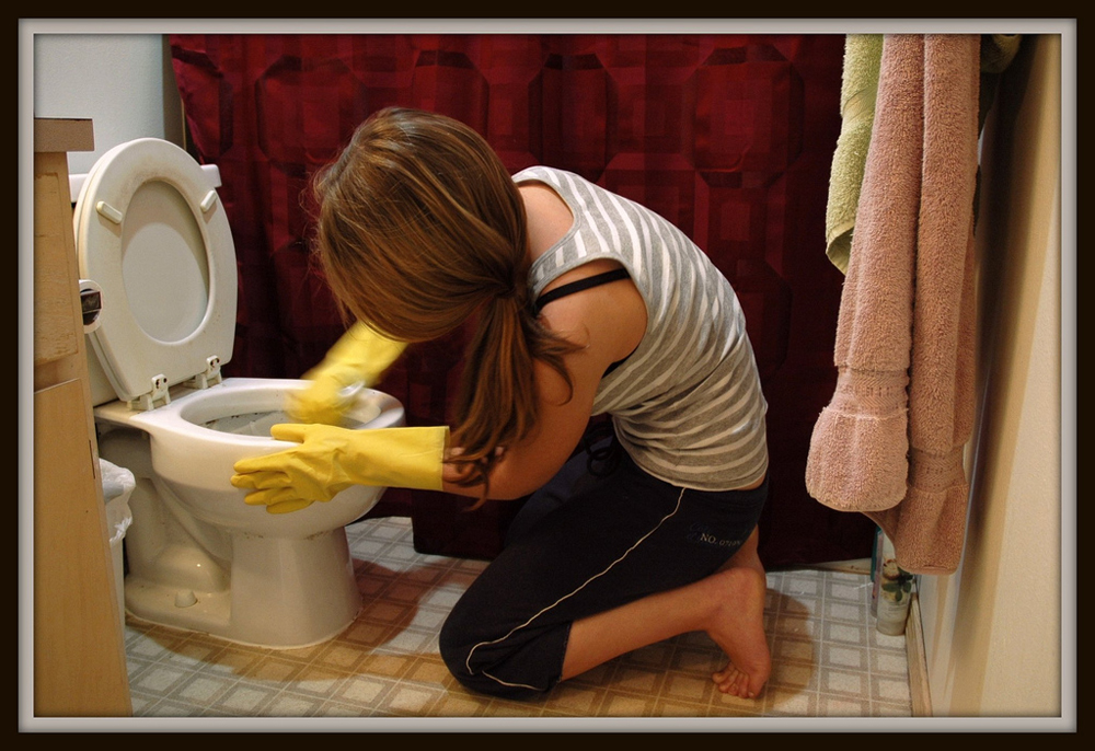 Photo of woman kneeling on bathroom floor scrubbing toilet.