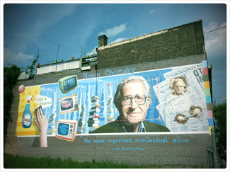 A mural on the side of a building. The mural includes Chomsky's face, along with some newspapers, televisions, and cleaning products. At the top of the mural, it reads “Noam Chomsky.” At the bottom of the mural, it reads “the most important intellectual alive.”