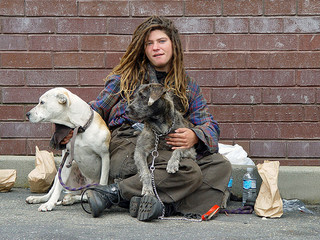 Photo of homeless woman sitting on the ground in front of a brick wall with two dogs.