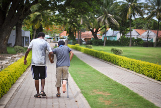 Photo of two men walking down a path. One is helping the other who is using a crutch.