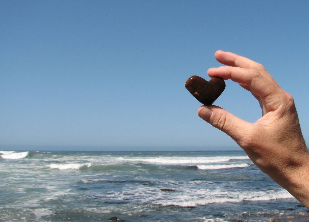 A person holding a heart shaped rock over looking an ocean