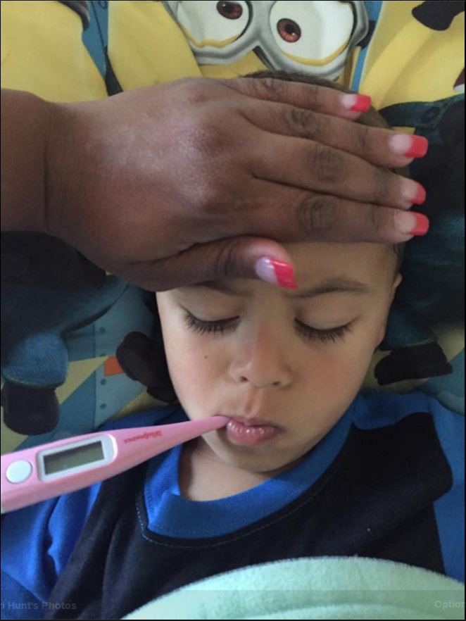 A sick child with a thermometer in their mouth and an adult's hand over their head checking their temperature.