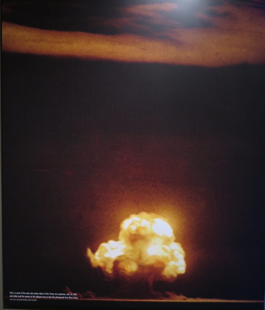 Colored photograph of Trinity nuclear explosion shaped like multiple clustered mushrooms