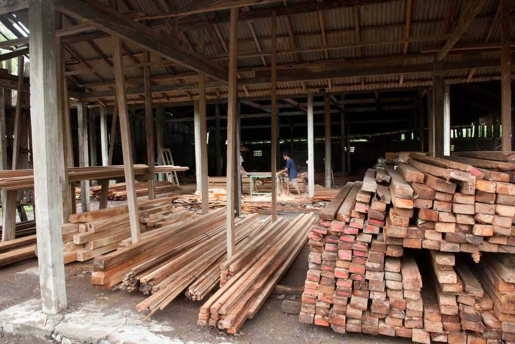 Pieces of lumber stacked with two people cutting wood in the background.