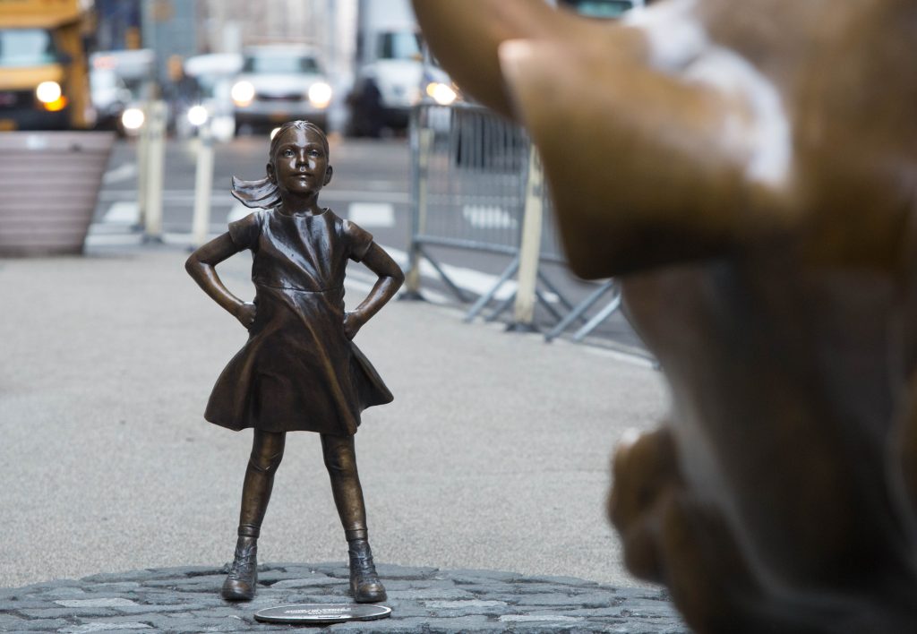 Bronze statute of a girl with her hands on her hips facing a bronze bull statute.