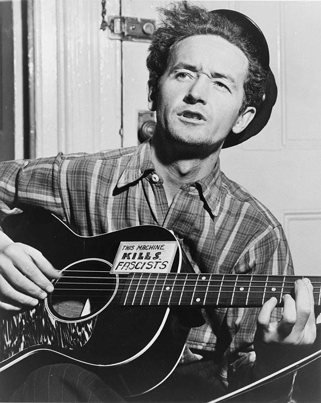 Woody Guthrie singing and playing a guitar that reads "This machine kills fascists"
