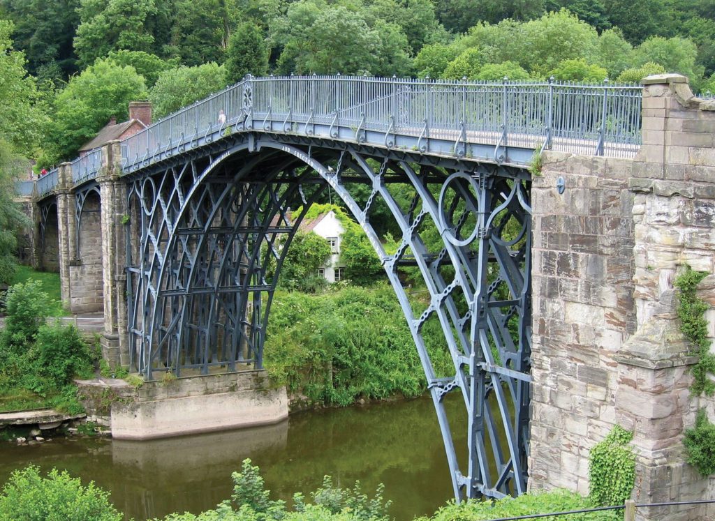 An image depicting a bridge made of iron and stone over a little river.