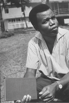 Photograph of Chinua Achebe, a dark skinned man sitting outside holding a closed book.