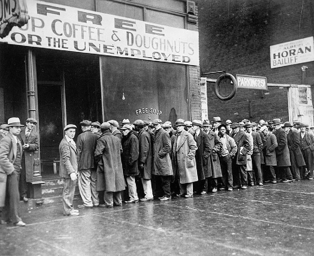"Men are lined up outside a building that has a sign that reads “Free Cup Coffee and Doughnuts for the Unemployed.”"