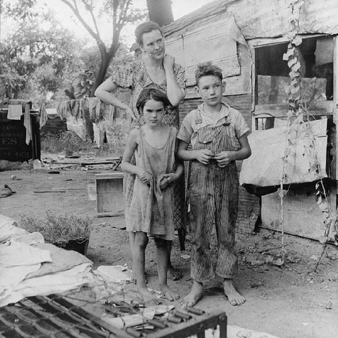 The photograph captures a woman and two children. They are barefoot and their clothes look dirty. Trash is visible nearby..