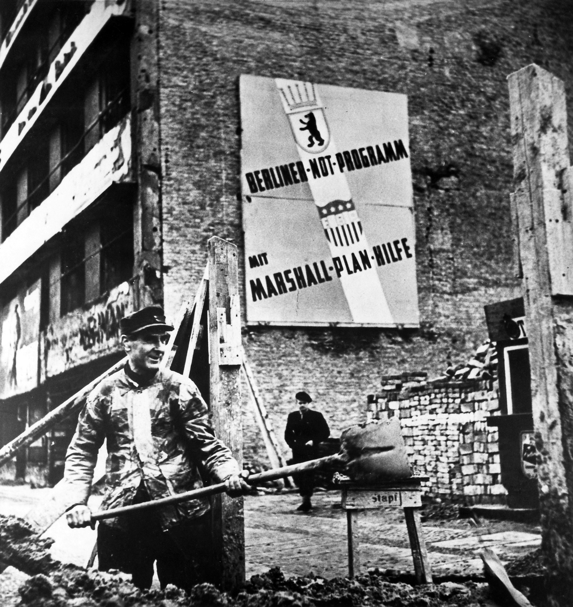 Photo of man with shovel working to build the Berlin Wall. There is a large sign a building behind the man that reads Emergency Program Berlin – with the help of the Marshall Plan"