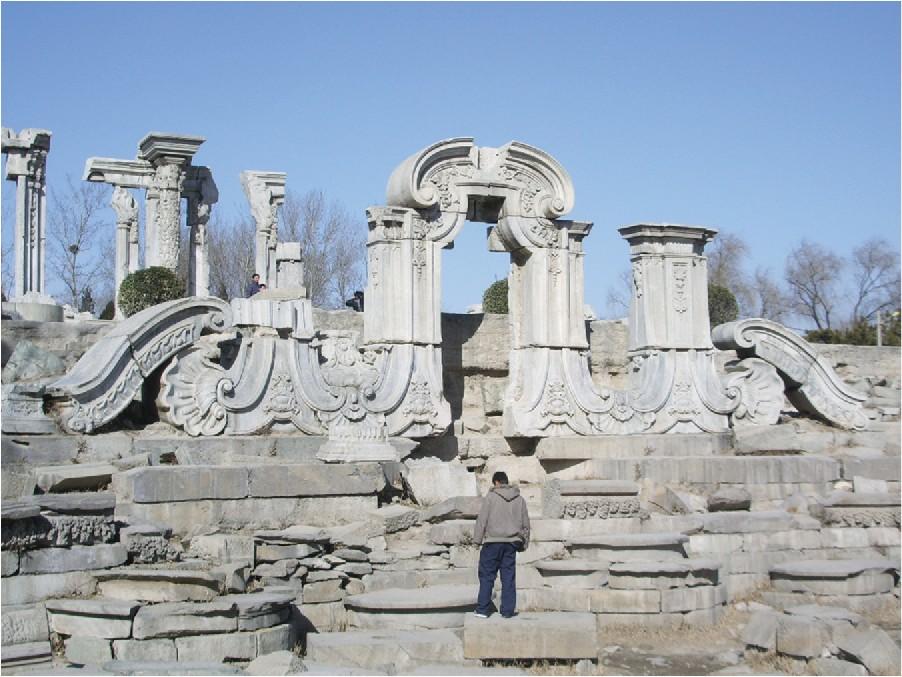 A photograph shows a very detailed white stone archway surrounded by collapsed and broken archway pieces. In the back left of the picture are five tall broken columns that are richly engraved. Trees run along the background.