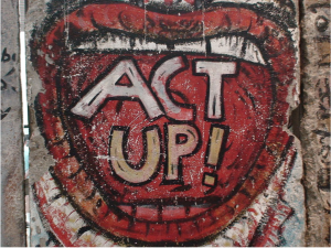 A picture of three sections of a concrete wall is shown. A mural of an open mouth is drawn on thelarger middle section. It has red lips, a red painted inside, and teeth, white on top and red at the bottom. The words “Act Up!” are painted inside the mouth, “act” in white and “up!” in tan. The side sections of the well have dark scribblings on them.