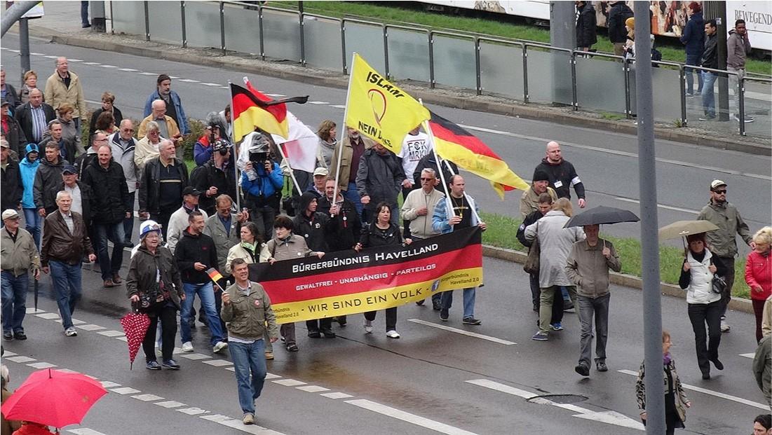 A picture of a large group of men and women walking on a wet street is shown. They wear pants, jackets, shoes, and hats. Four have umbrellas. Four people at the front of the group hold a large banner that is black, red, and yellow striped. The words “Burgerbundnis Havelland” are written in white in the top black strip, “Gewaltfrei – Unabhangig – Parteilos” are written in the middle red stripe in white and in black on the bottom yellow stripe are the words “-Wir Sind Ein Volk–” in the middle with “Burgerbundnis Havelland” in smaller black letters on both sides. A white “F” in a blue box is also seen at the right. Behind the banner four people carry large flags. Two flags are black, red, and yellow striped while one is white with some red and one is yellow with the words “Islam” and “Neinn” seen, with the rest obscured. In front of the banner twelve people are standing and walking about, some with umbrellas, some hugging, some talking. In the background of the picture, there is an opaque gate on the curb with grass behind and six people standing, watching, and walking. Parts of a white building can be seen behind the people.