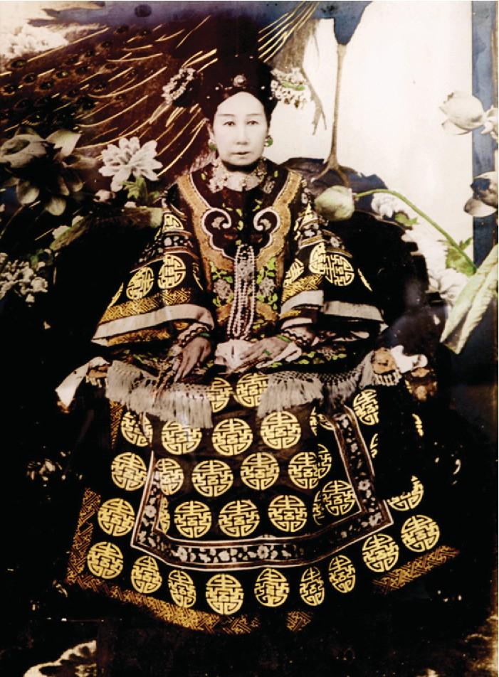 A photo of a woman with a very pale, white face. She wears a long dress decorated with yellow stenciled circles and ornate stitching with very large, flowy sleeves with tassels on the ends. She wears a gold decorated black hat on her head and large earrings. The background shows large flowers.