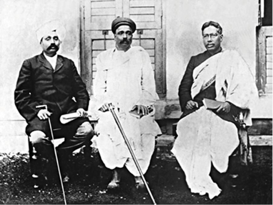 The photograph shows three men seated in chairs in front of a white house with shuttered doors. The man on the left wears a dark, long coat, pants, has a moustache, a white turban on his head and holds a cane in one hand a roll of papers in the other. The man in the middle wears a long white dhoti, has a moustache, a dark small cap, and holds a cane in one hand and a book in the other. The man on the right wears a dark coat and a large white cloth draped over his shoulder and covering his legs. He holds a book in his hand and wears glasses.