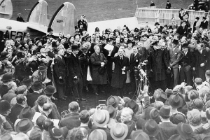 In this black and white picture, a man stands in the center of a large group of people, waving a piece of paper. A group surrounds him in a circle. Most of the crowd is men in suits and long coats and hats, with some women interspersed, in dresses and some with hats. There are microphones placed in front of the man, and there is the back of a plane visible in the back left of the picture.