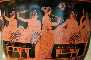 Image of a symposium on a Greek vase men are lying on couches and a woman plays