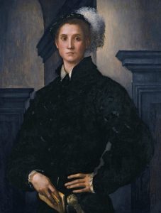 young man stands with one hand on his hip. He is dressed in black renaissance-era clothing and holds a book.