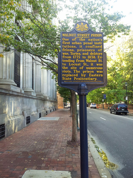 Historical marker on a street reading: Walnut Street Prison. One of the nation's first urban penal institutions, it confined felons, prisoners of war, Tories, and debtors from 1775 to 1838. Extending from Walnut St. to Locust St., it was the site of numerous riots. The prison was replaced by Eastern State Penitentiary.