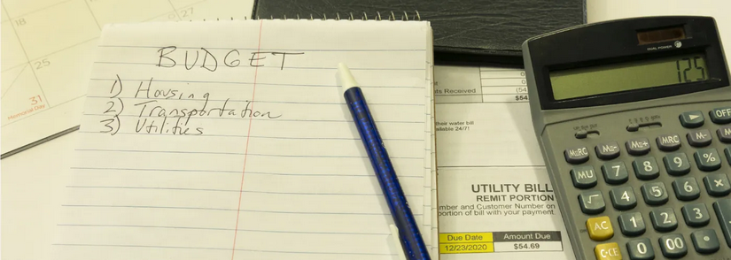 A photo of a calculator, a utility bill, a calendar, a notepad, and a pen. The notepad says BUDGET across the top with a numbered list: Housing, Transportation, and Utilities.