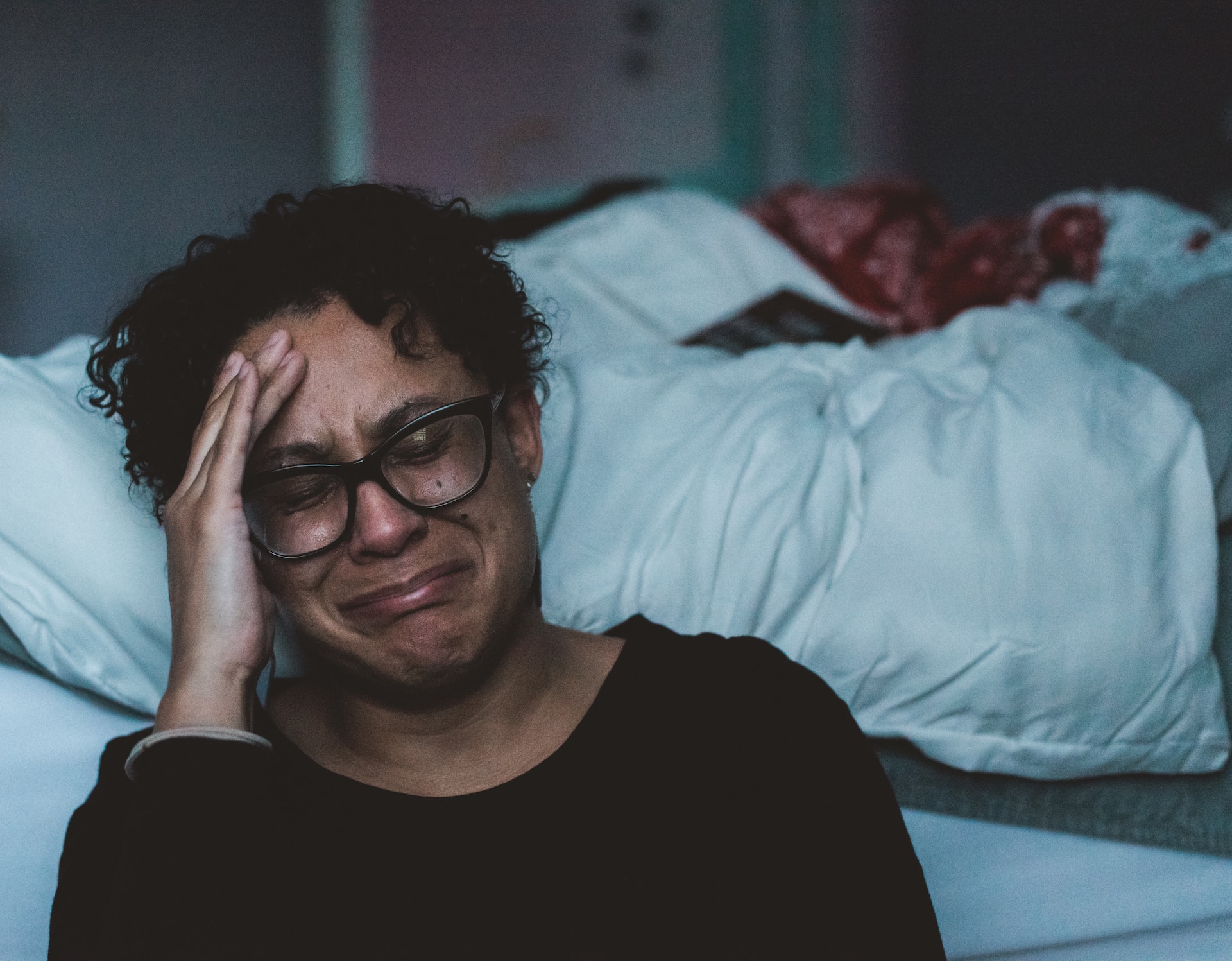 Photo shows a crying woman sitting beside a bed.