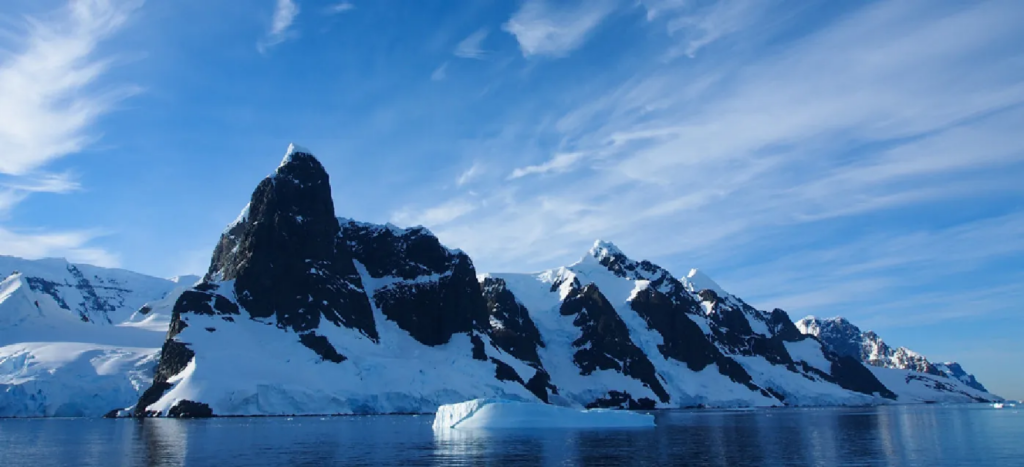 Picture of Antartica showing water, mountain, and iceberg peaks under a bright blue sky.