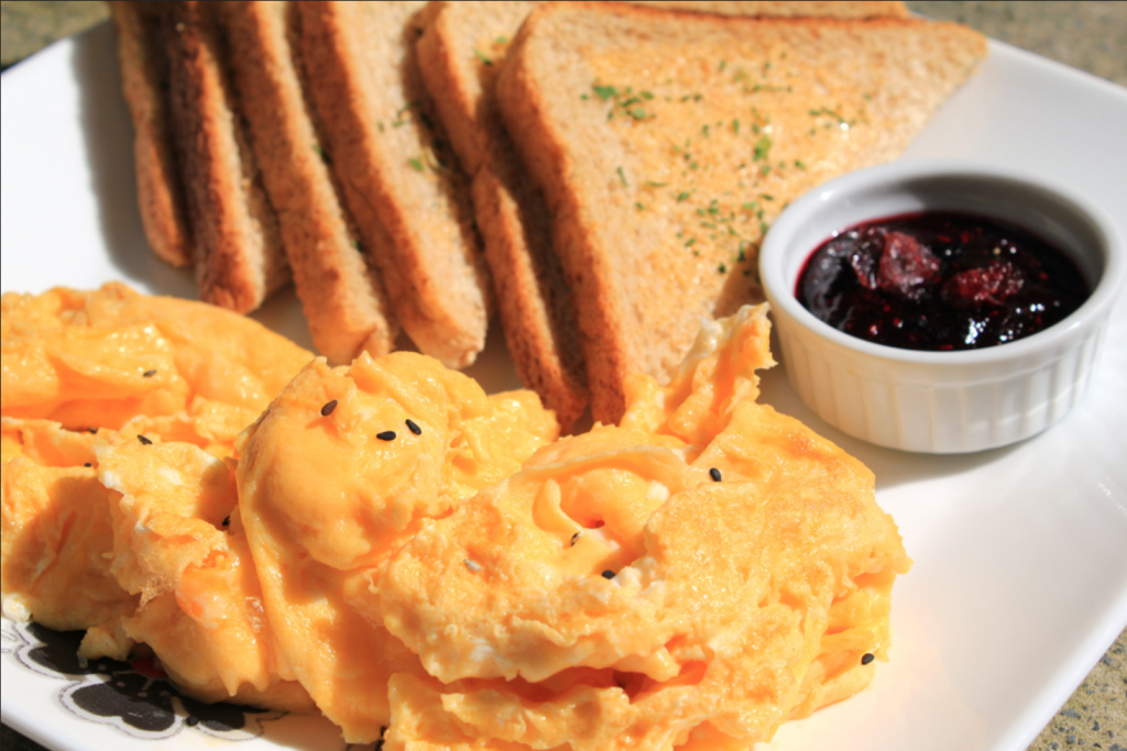 This is a photo of a plate with a large pile of eggs in the foreground and six slices of toast in the background. There is a small dish of red jam sitting near the toast on the plate.