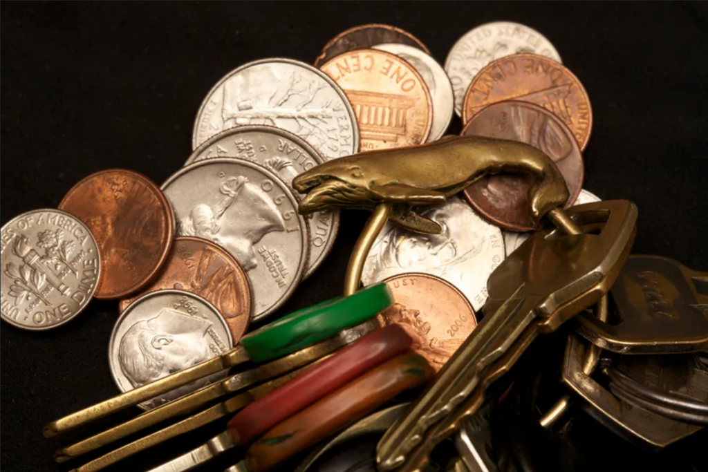 This is a photo of change and a set of keys in a pile. There appear to be five pennies, three quarters, four dimes, and two nickels. The key ring has a bronze whale on it and holds eleven keys.