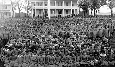 A photograph shows a large, posed group of Native American children at a school. The girls sit in the front in collared dresses. The boys stand at the back in button-down shirts and slacks.