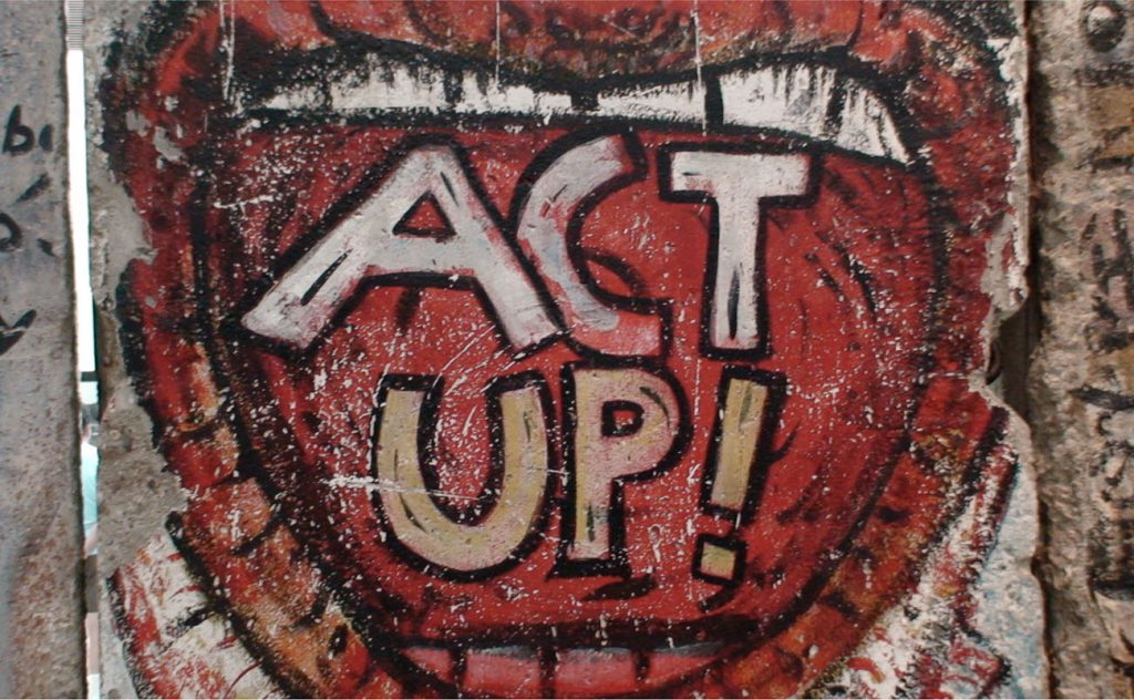 A panel of graffiti on the Berlin Wall shows a wide-open mouth, within which are the words “ACT UP!”